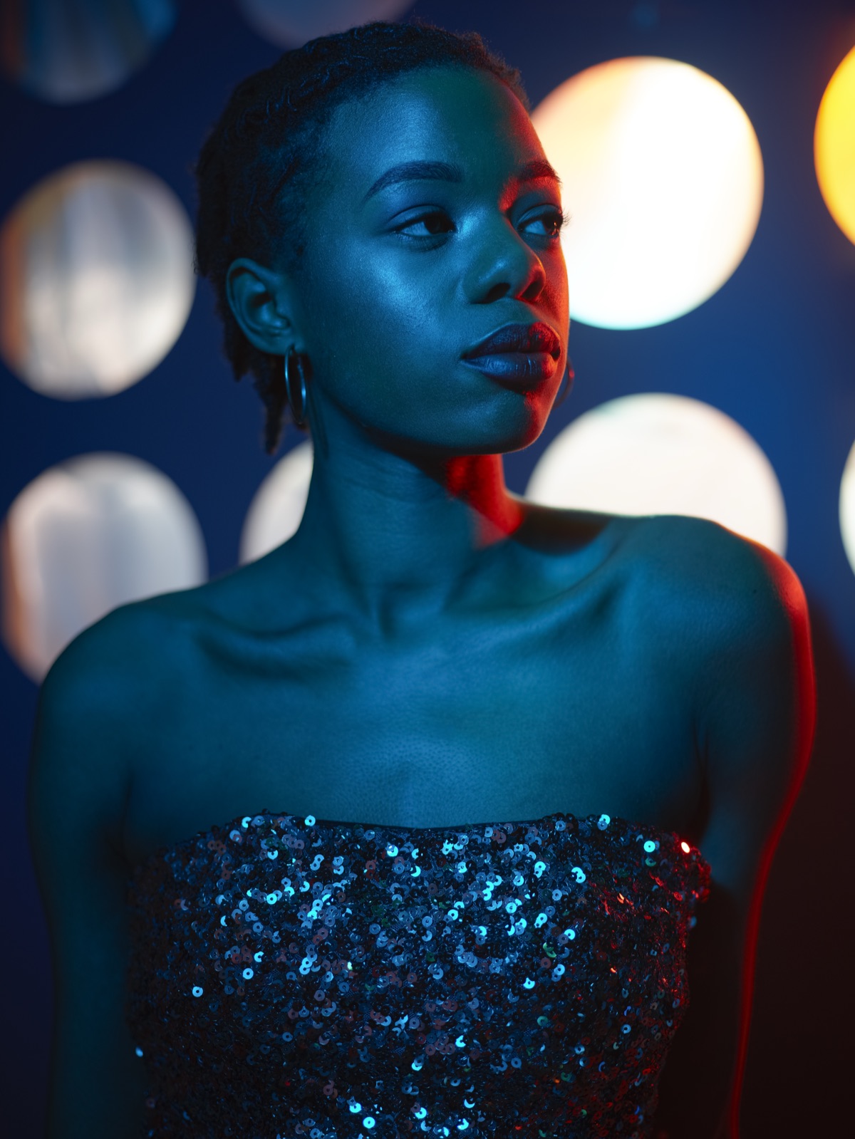 Portrait of a woman in a sequinned dress lit in blue and red against bokeh lights.