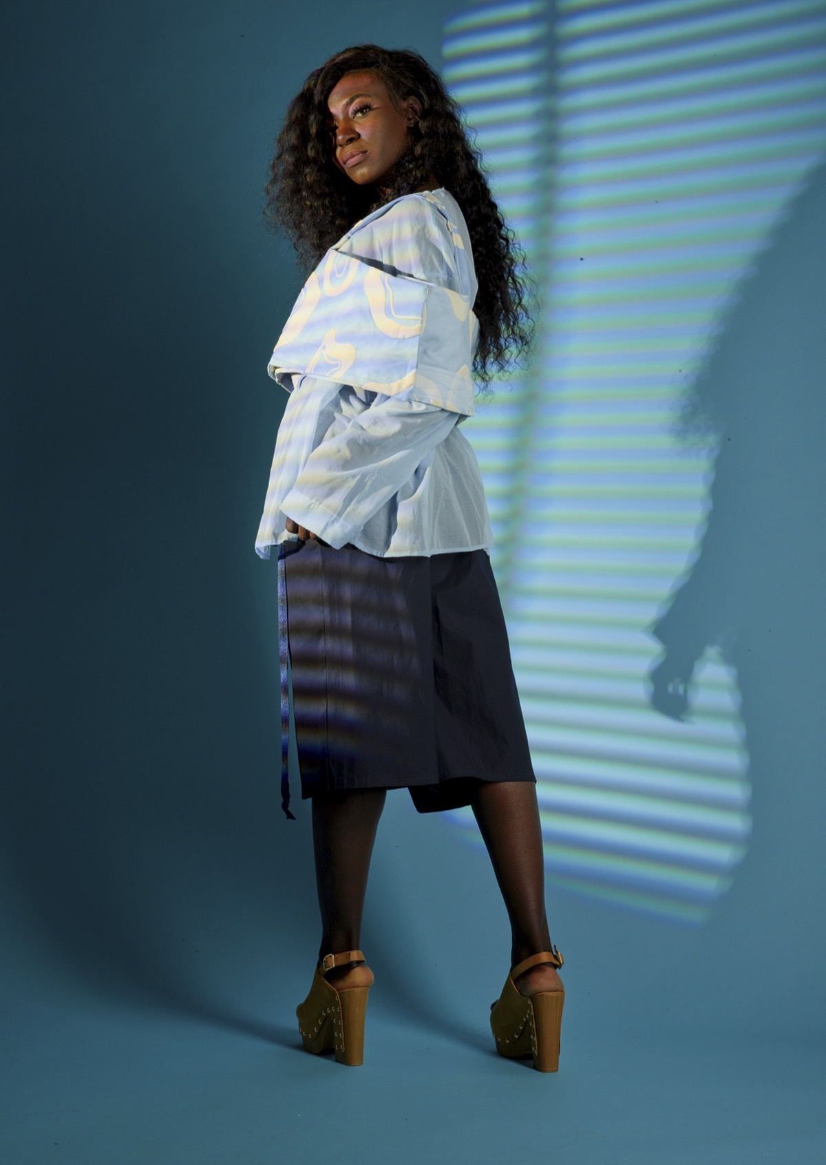 A model in a pale patterned jacket and dark skirt against a teal wall striped with projected light.