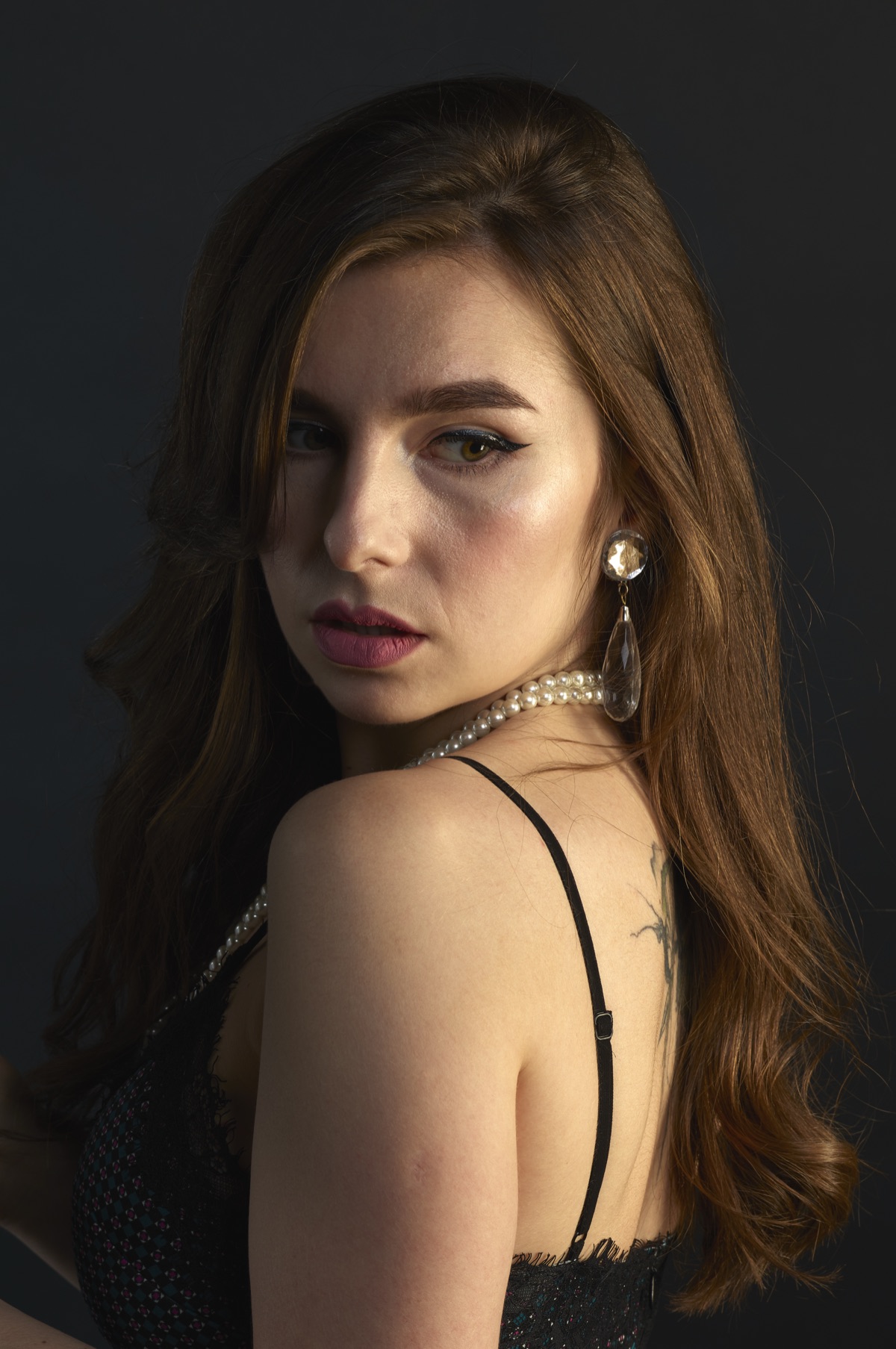 Portrait of a woman looking over her shoulder wearing pearl jewellery against a dark backdrop.