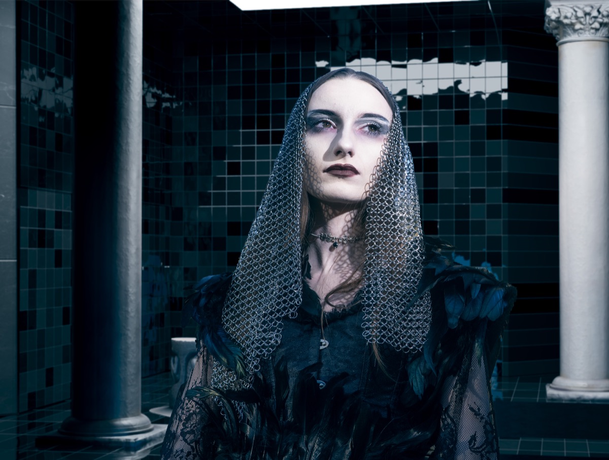A model in a chainmail veil and black feathered coat in a dark tiled room with stone columns.