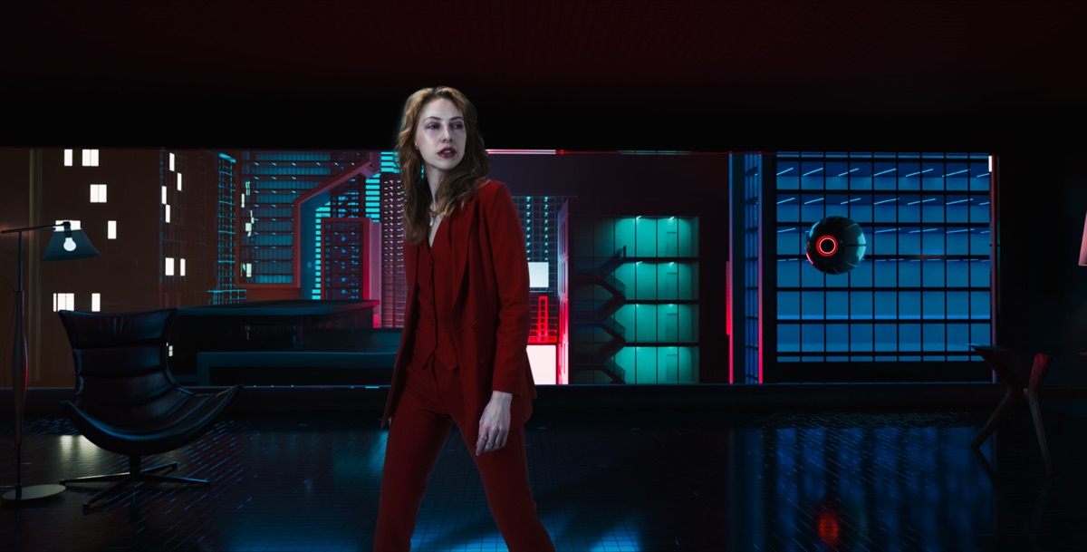 A woman in a red suit standing in a dark room lit by teal and red screens.